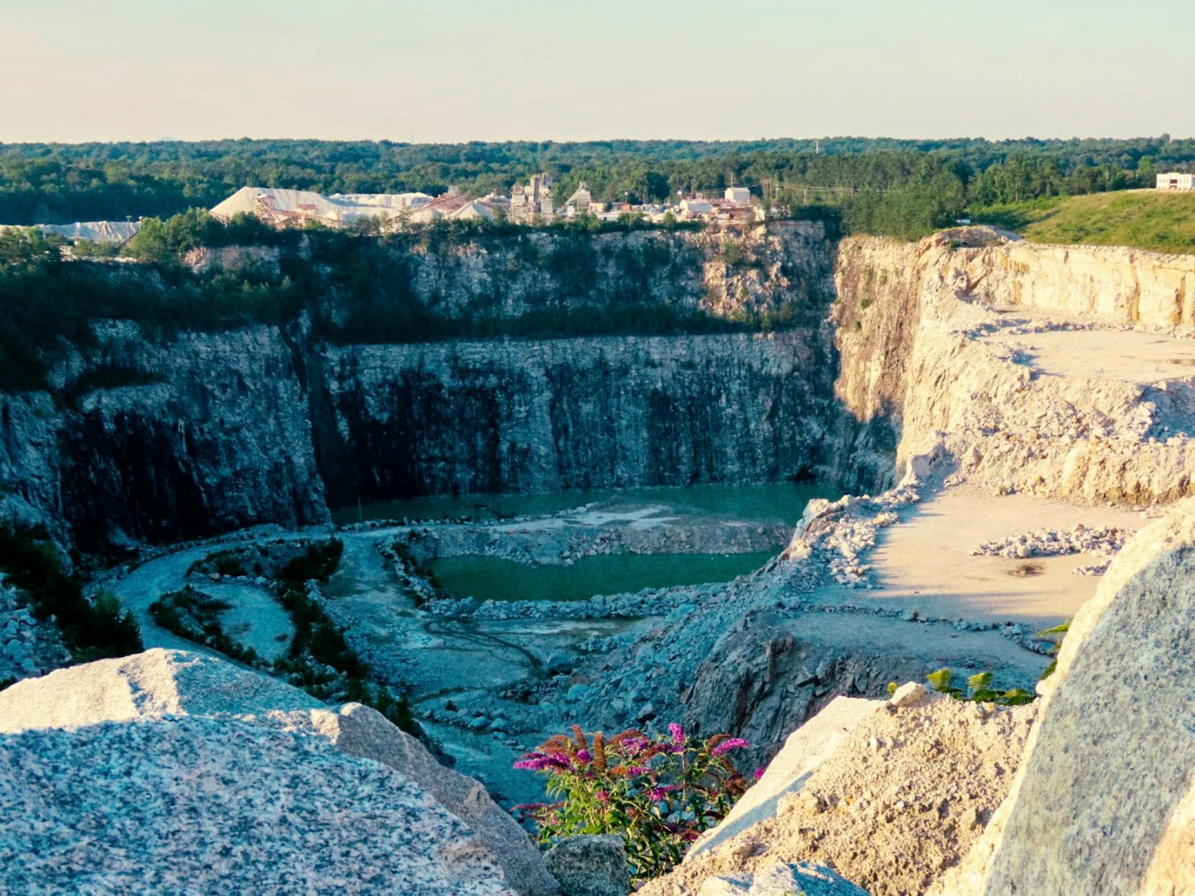 Explore the sunlit expanse of a large quarry with layered rock formations and distant industrial backdrop.