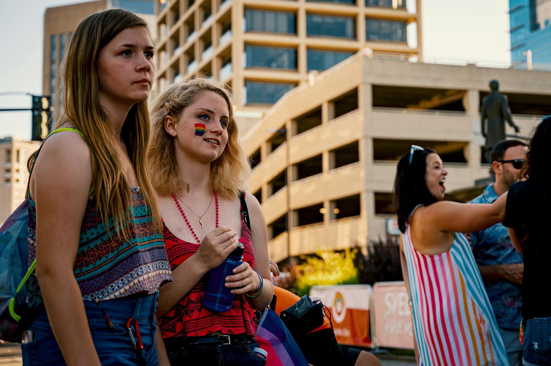 Two young women at an outdoor pride festival with urban buildings in the background.