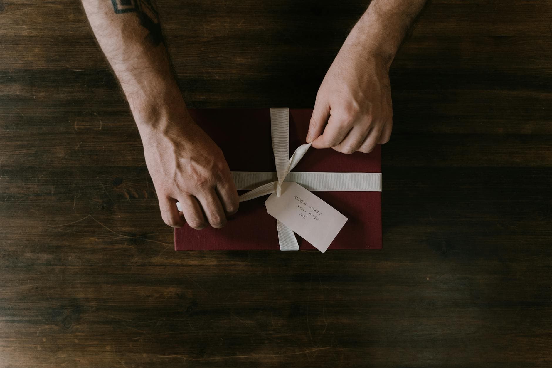Hands wrapping a red gift box with a white ribbon on a wooden surface, ideal for holiday or celebration themes.